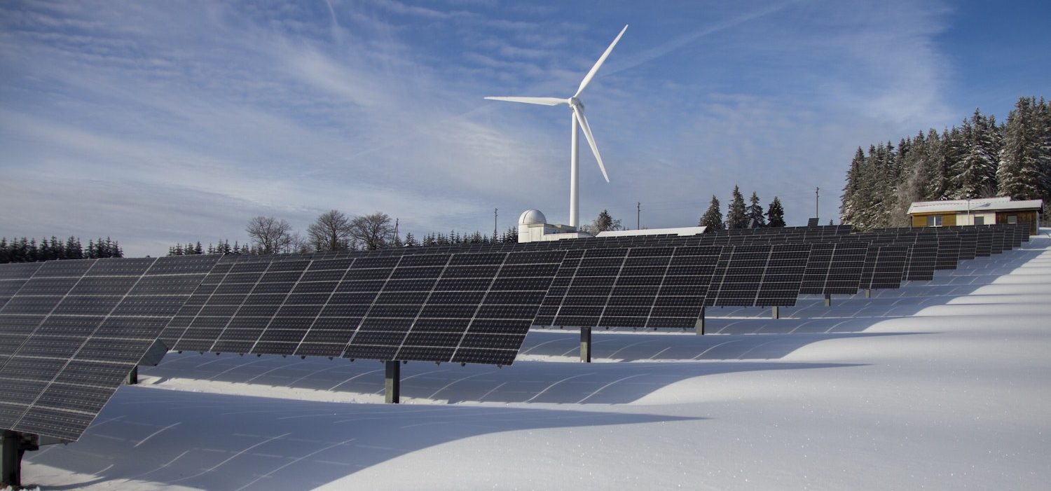 Solar panels and windmill in snowy field with farm in background.